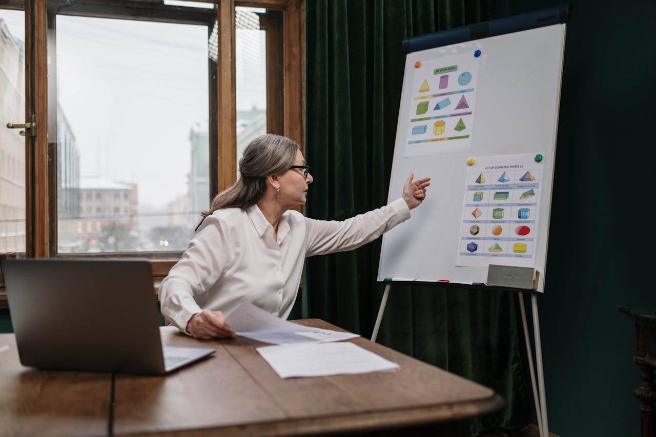 Senior woman conducting an online geometry lesson, highlighting various shapes on a chart with a laptop.