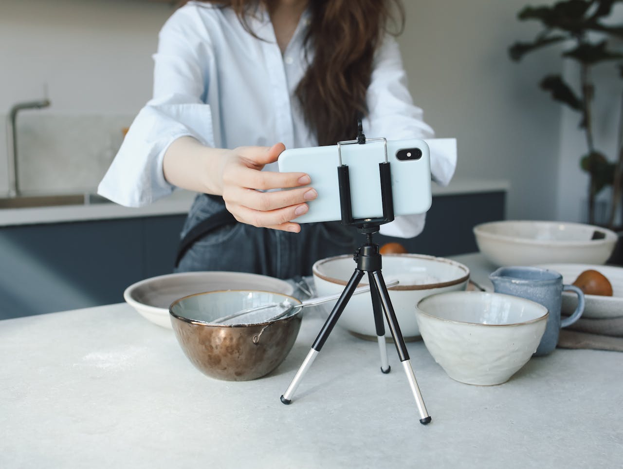 Woman using a smartphone on a tripod for vlogging in the kitchen with bowls.